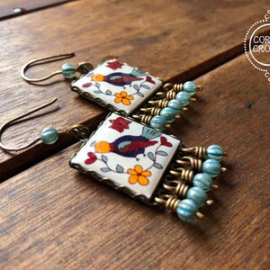 May include: Square earrings with a bird and floral design on a white background. The earrings have a bronze-colored frame and are accented with blue beads. The earrings are on a wooden surface.