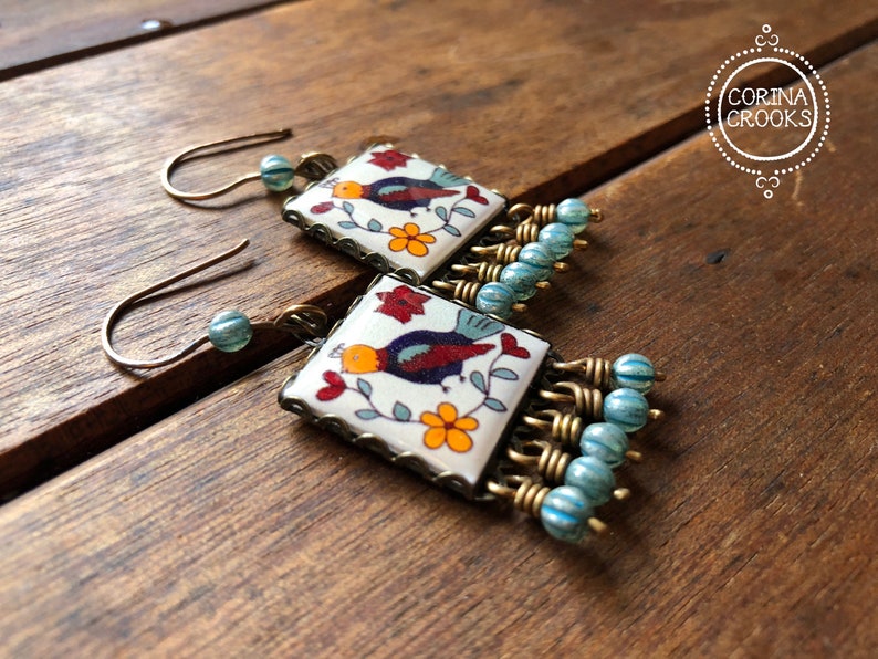 May include: Square earrings with a bird and floral design on a white background. The earrings have a bronze-colored frame and are accented with blue beads. The earrings are on a wooden surface.