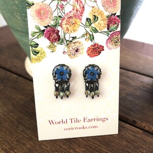 May include: A pair of World Tile Earrings with a floral design. The earrings have a blue flower center with a black frame and dangling beads. The earrings are displayed on a card with floral artwork and the website coricrooks.com.