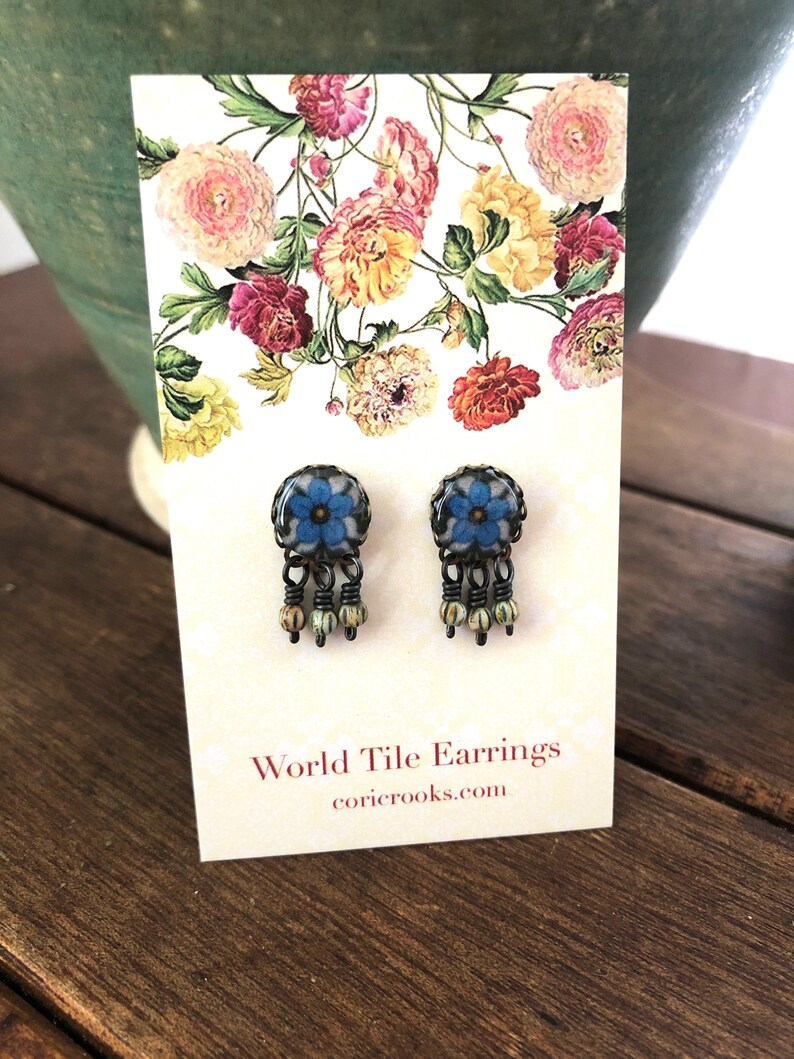 May include: A pair of World Tile Earrings with a floral design. The earrings have a blue flower center with a black frame and dangling beads. The earrings are displayed on a card with floral artwork and the website coricrooks.com.