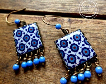 Puede incluir: Un par de pendientes cuadrados con un estampado floral blanco, azul y rojo. Los pendientes tienen un marco de color bronce y están adornados con cuentas azules. Se encuentran sobre una superficie de madera.