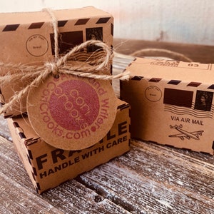 May include: Three small, brown cardboard boxes tied with twine, featuring airmail graphics and the text "VIA AIR MAIL." One box has a tag with "coricrooks.com" printed on it. The boxes are on a rustic wooden surface.