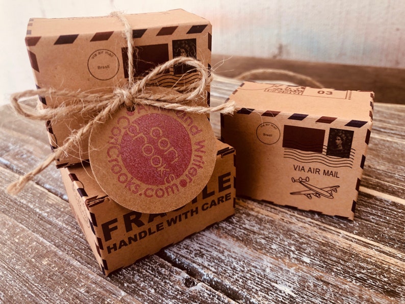 May include: Three small, brown cardboard boxes tied with twine, featuring airmail graphics and the text "VIA AIR MAIL." One box has a tag with "coricrooks.com" printed on it. The boxes are on a rustic wooden surface.