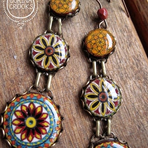 May include: A pair of handmade earrings with a series of circular decorative elements. The earrings display a floral design with yellow, red, and blue accents on a white background. They are linked by brass-coloured metal and have a hook closure.