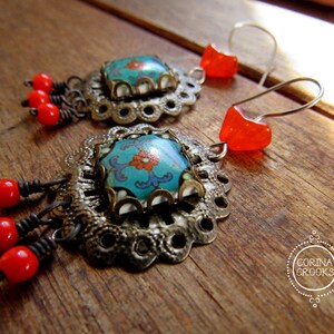 May include: A pair of ornate earrings featuring turquoise and orange floral designs. The earrings have silver-toned filigree settings, red bead accents, and orange heart-shaped ear wires. The earrings are displayed on a wooden surface.