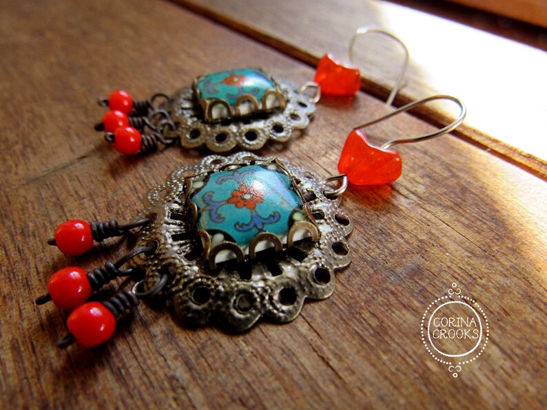 May include: A pair of ornate earrings featuring turquoise and orange floral designs. The earrings have silver-toned filigree settings, red bead accents, and orange heart-shaped ear wires. The earrings are displayed on a wooden surface.