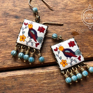 May include: Square earrings with a bird design. The earrings feature a white background with a colorful bird, flowers, and red accents. They have a brass frame and dangling blue beads. The earrings are displayed on a wooden surface.