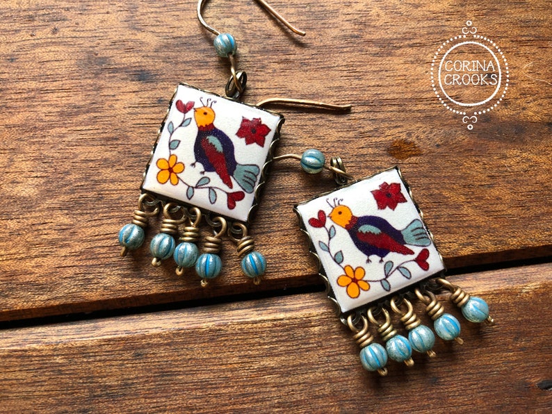 May include: Square earrings with a bird design. The earrings feature a white background with a colorful bird, flowers, and red accents. They have a brass frame and dangling blue beads. The earrings are displayed on a wooden surface.