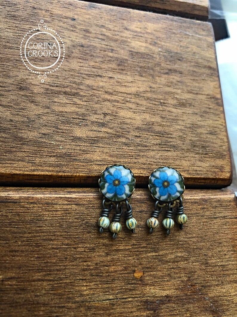 May include: A pair of floral stud earrings. Each earring features a blue flower design with green accents, set in a round, antique-style metal frame. Dangling below are three small, striped beads. The earrings rest on a wooden surface.