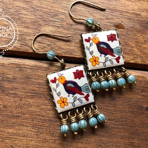 May include: Square earrings with a bird design. The earrings feature a white background with a colorful bird, red flowers, and yellow flowers. The earrings have a gold-colored metal frame and blue beaded fringe. The earrings are on a wooden surface.