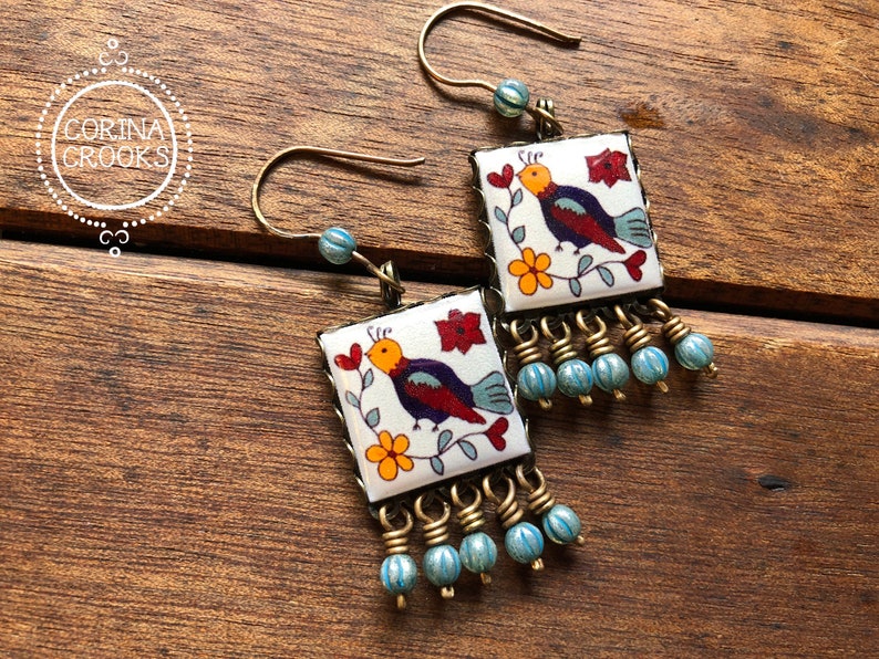 May include: Square earrings with a bird design. The earrings feature a white background with a colorful bird, red flowers, and yellow flowers. The earrings have a gold-colored metal frame and blue beaded fringe. The earrings are on a wooden surface.