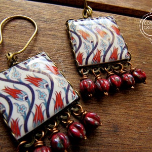 Può includere: Un paio di orecchini con un design a piastrella quadrata che presenta uno sfondo bianco con motivi floreali rossi, blu e neri. Le piastrelle sono accentate da una frangia di perline rosse.