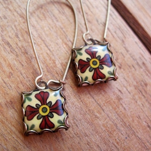 May include: A pair of square earrings with a floral design. The earrings feature a cream-colored background with a red and yellow flower design. The edges are trimmed with a gold-colored metal. The earrings are on gold-colored ear wires.