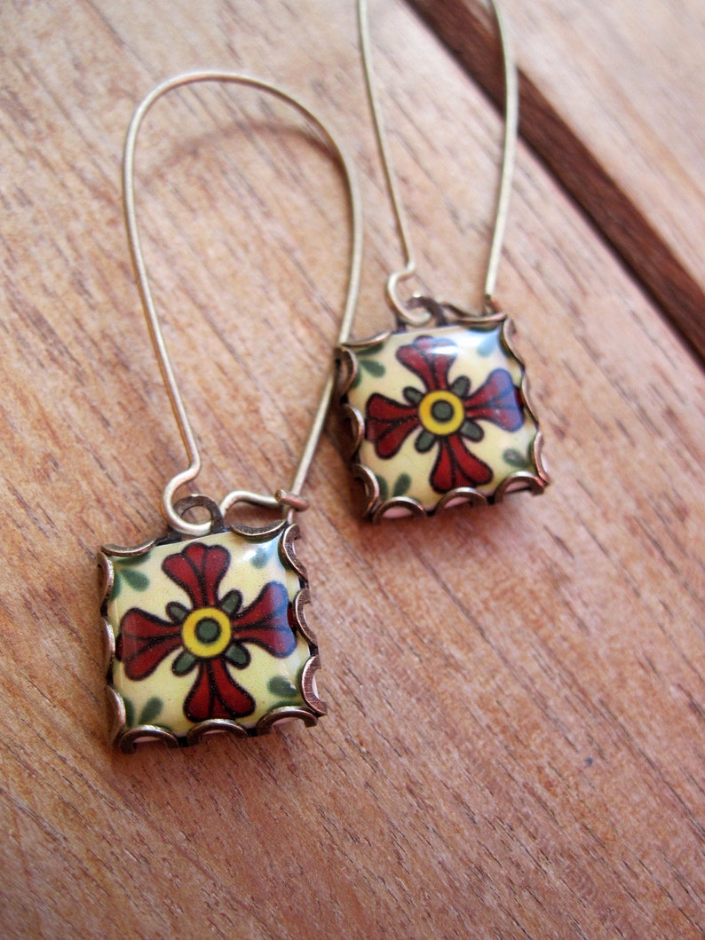 May include: A pair of square earrings with a floral design. The earrings feature a cream-colored background with a red and yellow flower design. The edges are trimmed with a gold-colored metal. The earrings are on gold-colored ear wires.