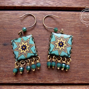 May include: A pair of square earrings with a turquoise and yellow starburst design. The earrings have a brass-colored frame and dangle with green beads. The earrings are on a wooden surface. The text "CORINA CROOKS" is in a circle.