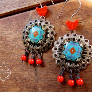 May include: A pair of ornate earrings featuring a turquoise and orange floral design at the center, surrounded by a silver filigree. Red beads dangle below, and a red heart-shaped bead tops each earring. The earrings are displayed on a wooden surface.