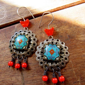 May include: A pair of ornate earrings with a floral design. Each earring features a turquoise and orange floral center, surrounded by a silver-colored filigree. Red beads dangle from the bottom, and a red heart-shaped bead sits at the top. The earrings are displayed on a wooden surface.