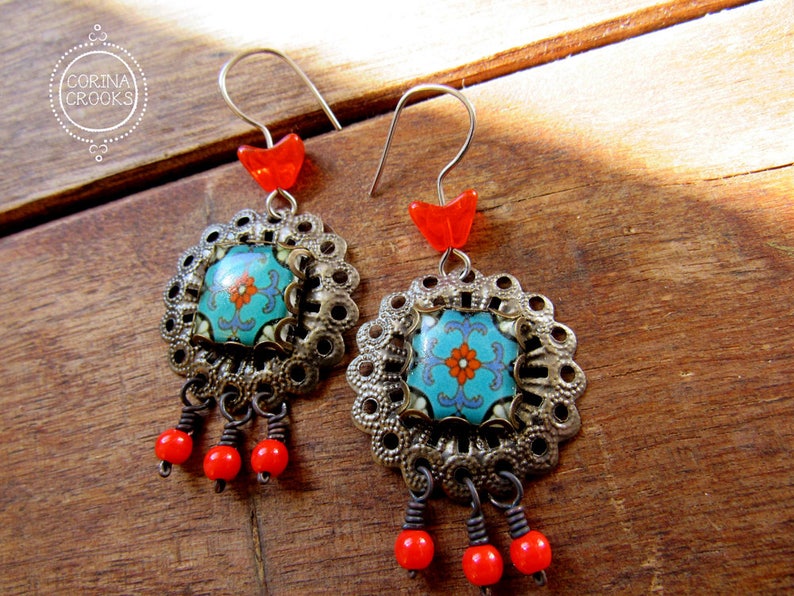 May include: A pair of ornate earrings with a floral design. Each earring features a turquoise and orange floral center, surrounded by a silver-colored filigree. Red beads dangle from the bottom, and a red heart-shaped bead sits at the top. The earrings are displayed on a wooden surface.