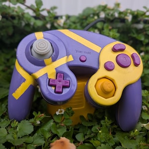 Pode incluir: Um comando de videojogos roxo com detalhes pintados em amarelo, um manípulo analógico cinzento, um D-pad roxo e botões de ação roxos com as letras A, B, X e Y. O comando inclui um pequeno botão Start/Pause roxo no centro da estrutura.