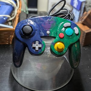 May include: A blue, green, and purple GameCube controller with a clear plastic base. The controller has a white plus button, a yellow c-stick, and a blue, green, and purple paint job.