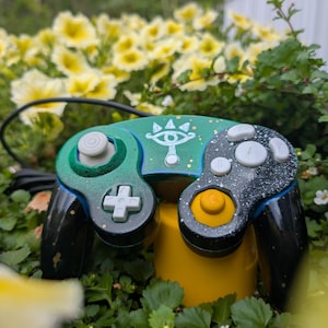 May include: A custom video game controller featuring a gradient finish transitioning from green to black with gold speckles. The center displays a white eye symbol. It includes a white D-pad, white buttons, a yellow analog stick, and a black cord.