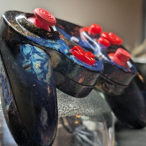 May include: A video game controller featuring a dark, glossy finish with swirling blue and gold nebula-like patterns. The device includes bright red analog sticks, a red directional pad, and red action buttons, all set against the dark, textured shell.