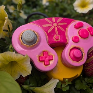 May include: A custom video game controller featuring a bright pink and magenta color scheme with hand-painted gold accents. The controller includes a gray analog stick, a pink directional pad, pink action buttons, and a gold sun-like design.