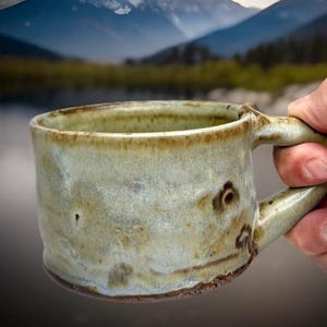 May include: A handmade ceramic mug with a rustic, textured appearance. The mug has a light green and brown glaze, with a rounded body and a sturdy handle. The mug is held in front of a scenic mountain and lake background.
