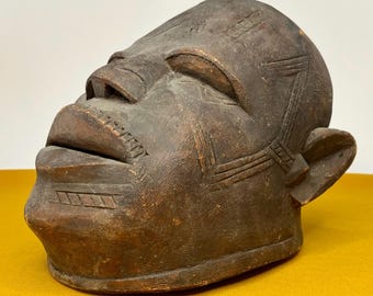 May include: A dark brown wooden mask depicting a human face with carved details. The mask has a closed mouth and eyes, and geometric patterns are carved into the forehead and sides.