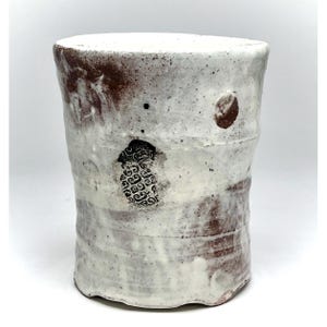 May include: A handmade ceramic tumbler with a textured surface. The cup features a white glaze with brown accents and a decorative patterned design. The design is a unique piece of pottery, perfect for enjoying beverages.