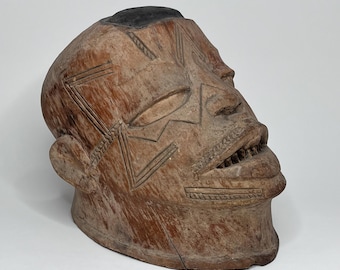 May include: A wooden mask carved in the shape of a human head. The mask has a dark brown color and features a carved mouth with teeth, eyes, and a nose. The mask is decorated with geometric patterns carved into the surface.