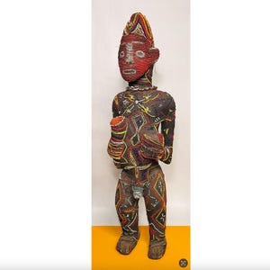 May include: A beaded figure with a red face and elongated head. The figure is adorned with intricate beadwork in various colors, including red, yellow, and black. It holds objects in its hands and has a necklace. The figure stands on a yellow surface.