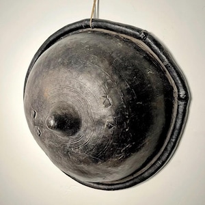 Puede incluir: Un cuenco grande, redondo, de arcilla negra con un centro elevado y un borde de cuero. El cuenco está colgado de una cuerda.
