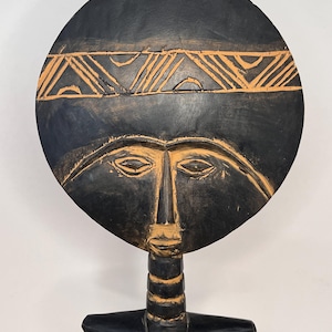 May include: A wooden sculpture of a stylized female figure with a circular head and geometric patterns. The sculpture is painted black with brown accents.