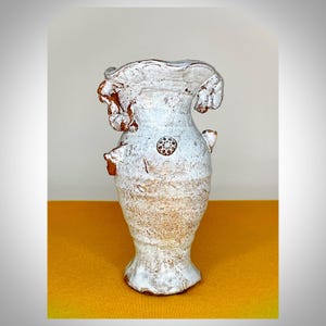 Peut inclure: Un vase en céramique fait à la main avec un glaçage blanc cassé texturé. Le vase présente un bord unique et froissé et de petites protubérances décoratives. Un motif circulaire est centré sur le corps. Le vase a une esthétique rustique et artisanale.