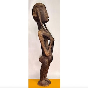 Puede incluir: Una estatua de madera tallada de una persona con una constitución esbelta, color marrón oscuro y una expresión serena. Los brazos de la figura están cruzados y tiene rasgos alargados. La estatua está de pie y parece ser una obra de arte tribal africana.