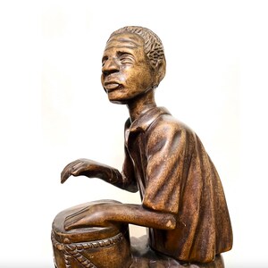 May include: A wooden sculpture of a person playing a drum. The figure is carved from brown wood and is seated, with hands positioned on the drum. The sculpture has detailed facial features and is set against a white background.