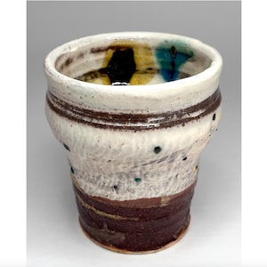 May include: Handmade ceramic cup with a unique design. The cup features a white exterior with brown bands and a dark brown base. It has colorful accents of black, yellow, and blue near the rim and small green and yellow dots on the body.