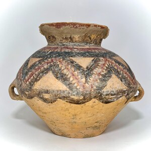 May include: A tan-colored, ancient-style pottery vessel with a wide body and a narrow neck. The vessel features a geometric design in black, red, and tan. Two small handles are on the sides. The rim is a darker shade of tan.