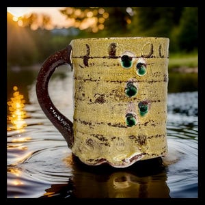 May include: A handmade ceramic mug with a textured, yellow-gold exterior and a dark brown handle. The mug features dark brown horizontal stripes and four green, jewel-like accents. The mug is sitting in water, reflecting the sunlight.