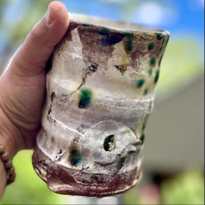 May include: Handmade ceramic tumbler with a unique, textured surface. The cup features a blend of colors, including white, brown, and green accents. The base is a deep reddish-brown. The design is organic and rustic, perfect for enjoying beverages.
