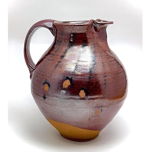 May include: A ceramic pitcher with a rounded body and a curved handle. The surface features a glossy, deep reddish-brown glaze with dark streaks and small, circular tan-colored spots. The base is unglazed, revealing a natural, matte tan clay texture.