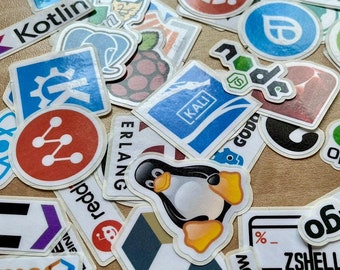 Op de afbeelding: Een verzameling diverse vinylstickers met verschillende logo's voor softwareontwikkeling, iconen van programmeertalen en emblemen van technologiemerken in uiteenlopende vormen, kleuren en maten, verspreid over een plat oppervlak.