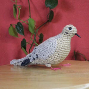 Puede incluir: Una paloma de ganchillo hecha a mano en tonos beige y gris, con manchas negras en las alas y la cola. El ave tiene un pico negro y patas de alambre rosa, sobre un fondo rojo con follaje verde.