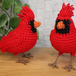May include: Two crocheted cardinal birds, one red with a black eye and orange beak, the other red with a black eye and orange beak, both with brown legs and feet.
