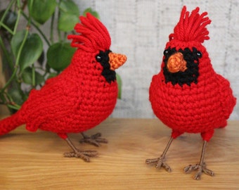 Puede incluir: Dos pájaros cardenal tejidos a crochet, uno rojo con un ojo negro y un pico naranja, el otro rojo con un ojo negro y un pico naranja, ambos con patas y pies marrones.