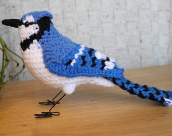Puede incluir: Un pájaro azul jay de crochet con detalles en blanco y negro. El pájaro tiene una cola larga y está de pie sobre una superficie de madera.