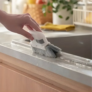 Könnte beinhalten: Eine weiße und graue Reinigungsbürste mit Griff und zwei Schrubbern. Die Bürste wird verwendet, um eine weiße Arbeitsplatte zu reinigen.