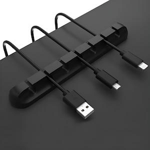 Puede incluir: Organizador de cables negro con seis ranuras para sujetar cables y alambres. El organizador está montado en una superficie de escritorio y tiene tres cables que pasan a través de él.