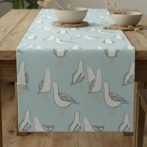 Peut inclure: Chemin de table bleu clair avec un motif répété de mouettes blanches avec des détails gris et jaunes. Le chemin de table est présenté sur une table en bois, ajoutant une touche de charme côtier à la salle à manger.